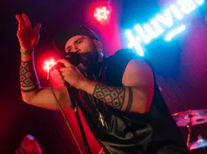 A singer with patterned arm tattoos performs on stage, holding a microphone under red lights, with a neon sign in the background.